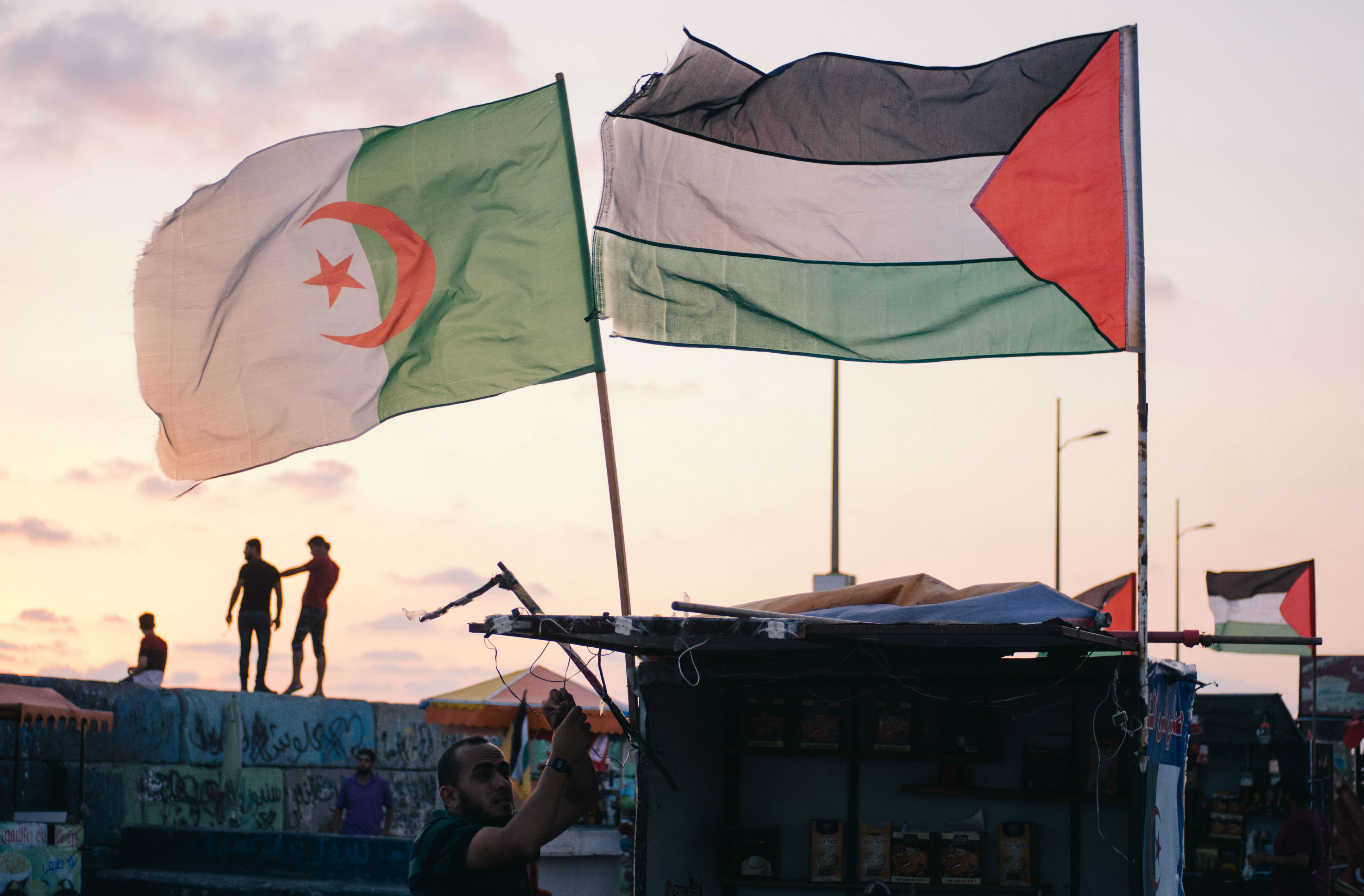 A vendor sets up his coffee stall decorated with the Algerian flag, catering for the families who whittle away the evening hours in the port. Palestinian youths climb up the breeze blocks behind him, posing for selfies against the sunset 