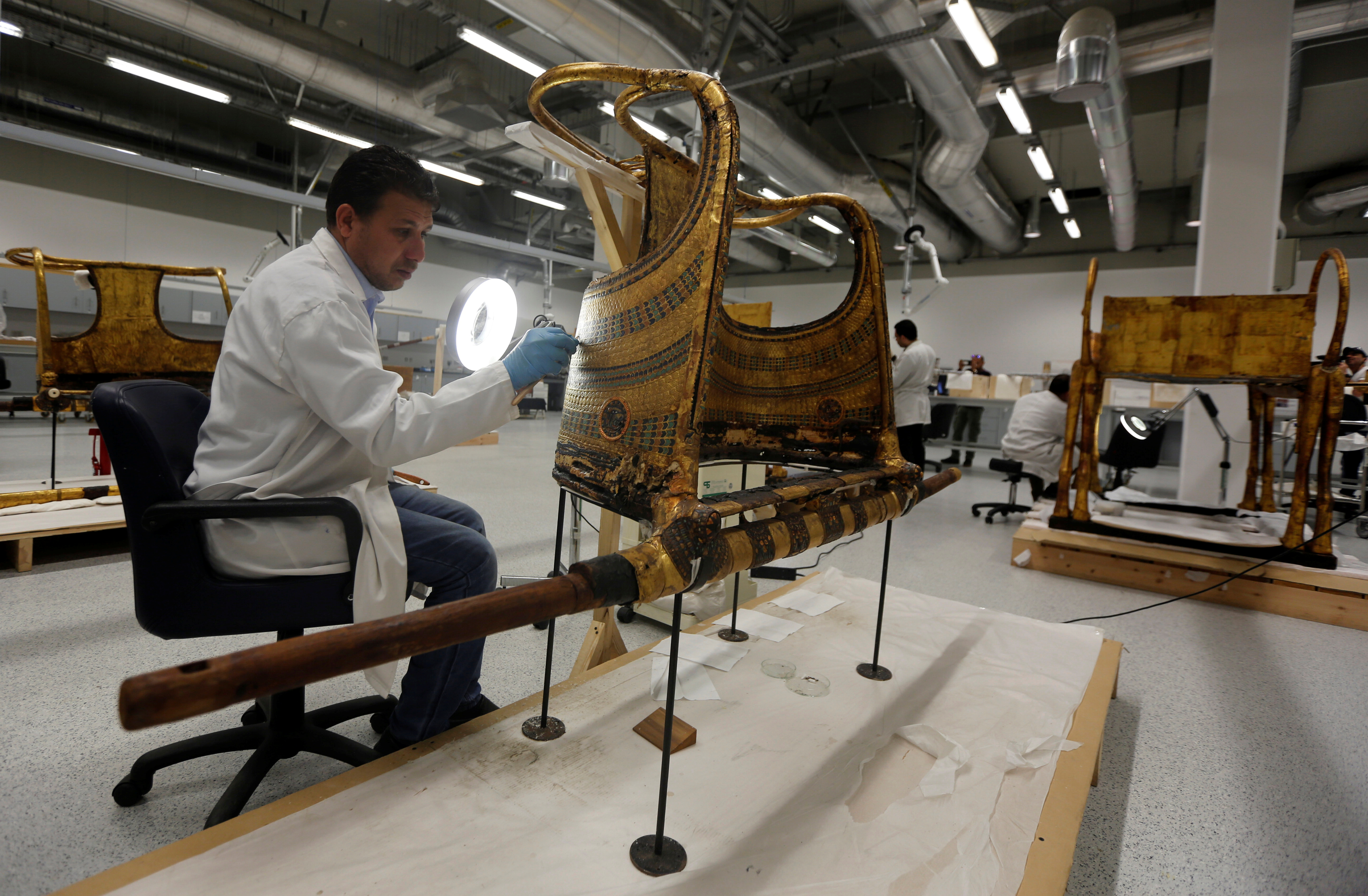 Here, an Egyptian archaeological technician renovates the golden war wheel which belonged to Tutankhamun: other treasures visible in the background include a gold pharonic bed to the right (Reuters)